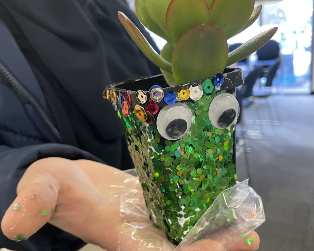 A kid holds a bedazzled pot plant in their hand (featuring googly eyes!)